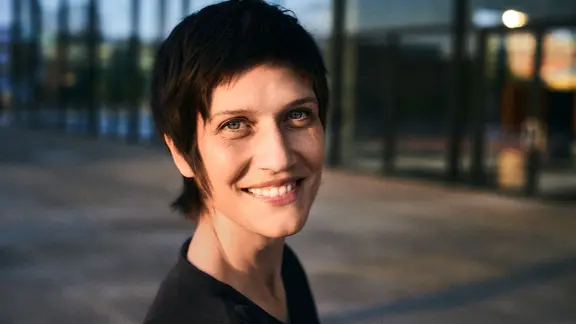 Alin Coen, eine junge Frau im schwarzen T-Shirt und mit kurzen Haaren, lächelt in die Kamera.