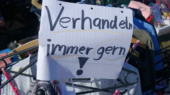 An einem Stand auf einem Flohmarkt hängt ein selbstgeschriebener Zettel mit der Aufschrift "Verhandeln immer gern!"