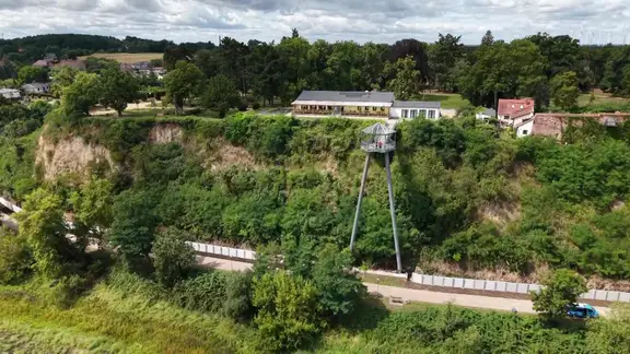 Ein begrünter Hang mit einem Aussichtsturm, am Fuße des Hangs eine graue Mauer.