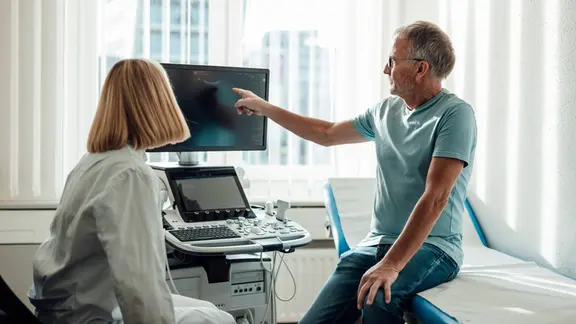 Patient zeigt auf Ultraschallmonitor und berät sich mit einem Arzt in einem Innenraum.
