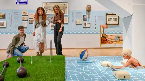Drei Frauen schauen sich eine Ausstellungsfläche an. Verschiedene Badeutensilien sind ausgestellt.