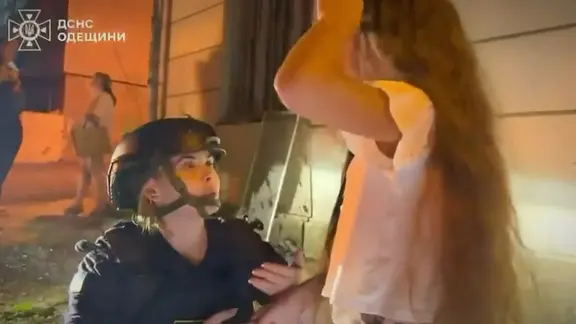 Frau mit Helm vom Katastrophenschutz hockt nachts vor einem Mädchen vor einem Gebäude.