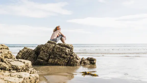 Eine Frau sitzt auf einem Felsen am Strand und hört etwas mit Kopfhörern.