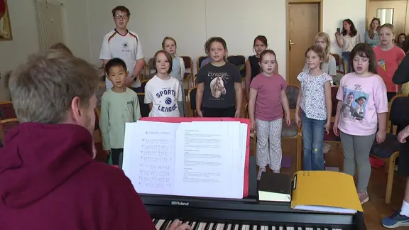 Kinder singen im Chor und werden von einem Klavier begleitet.