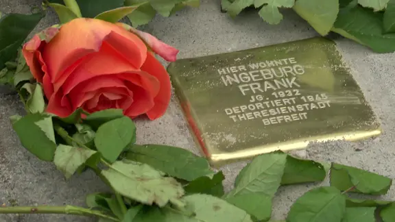 Ein goldfarbener so genannter "Stolperstein" mit dem eingravierten Namen Ingeburg Frank in einem Sandbett; daneben eine orangerote Rose