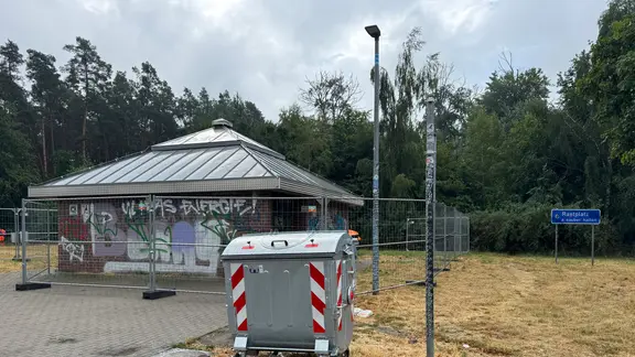 Ein vollgesprühtes und mit einem Bauzaun umschlossenes Toilettenhäuschen auf einem Rastplatz mit einer Mülltonne im Vordergrund.