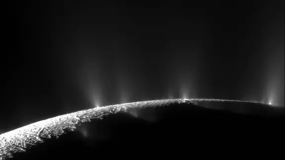 Schwarzweißaufnahme der Krümmung am Südpol von Saturnmond Enceladus mit Eiskruse: Entlang des hell erleuchteten Horizonts steigen mehrere Lichtstrahlen in die dunkle Atmosphäre auf.