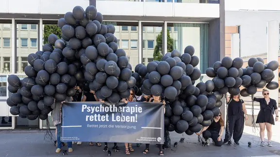 Bei einer Demonstration halten mehrere Menschen ein Banner mit der Aufschrift "Psychotherapie rettet Leben! Rettet jetzt die Versorgung" sowie viele schwarze Luftballons mit weißen Kreuzen hoch