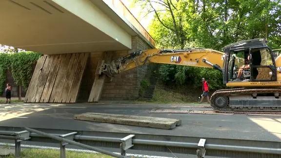 Ein Bagger stellt mit seinem Greifarm eine Stütze aus Holz oder Beton unter eine Brücke