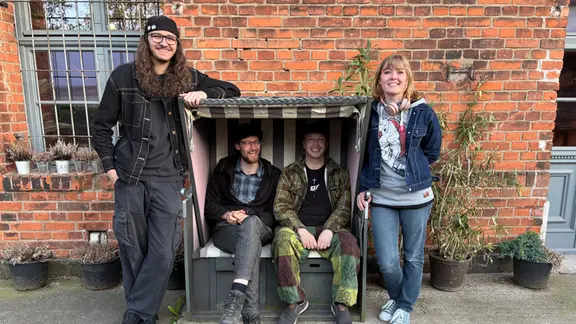 Gruppenfoto vor einer Backsteinwand eines alten Fabrikgebäudes: vier junge Menschen sitzen in oder stehen neben einem Strandkorb und lächeln in die Kamera