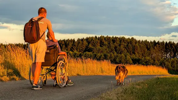 Eine Pflegekraft begleitet eine zu pflegende Person im Rollstuhl auf einem einsamen Weg in Richtung eines dunklen Horizonts.