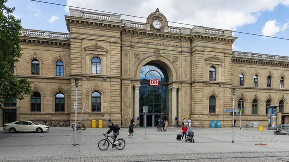 Blick auf den Haupteingang des Magdeburger Hauptbahnhofs, vor dem Reisende mit Koffern und Taschen herum laufen. 