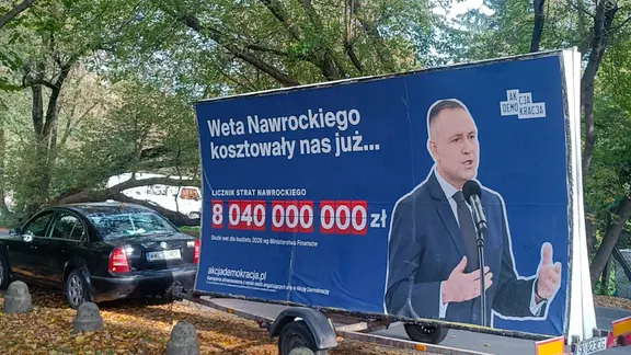 Schwarzer Pkw zieht einen Anhänger mit einem riesigen Plakat: "So viel haben uns bereits Nawrockis Vetos gekostet" (Polen)