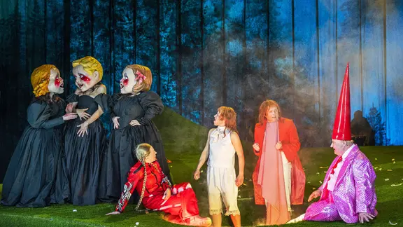 Szene aus dem Stück "Deutsche Märchen": Schauspielerinnen und Schauspieler stehen als Märchenfiguren verkleidet auf einer Bühne mit Waldszenerie im Hintergrund und Kunstrasen im Vordergrund. 
