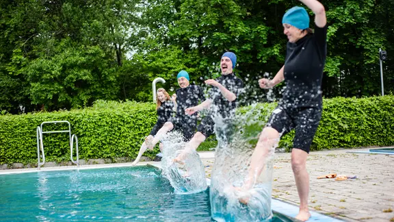 Vier Personen in Schwimmanzügen dippen performativ ihre Füße ins Wasser.