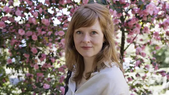 Anja Kampmann, eine Frau mit langen, rötlichen Haaren steht vor einem rosa blühenden Baum