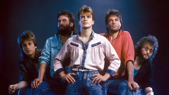 Die fünf Bandmitglieder von Karussell in V-Formation aufgeommen, mit 80er-Jahre-Blusen und Blue-Jeans.