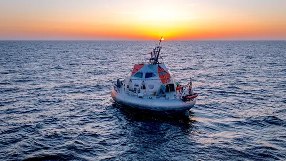 Rundliche Forschungsstation mit kuppelförmigem Aufbau fährt bei Sonnenuntergang über ruhige See, die Sonne steht tief über dem Horizont