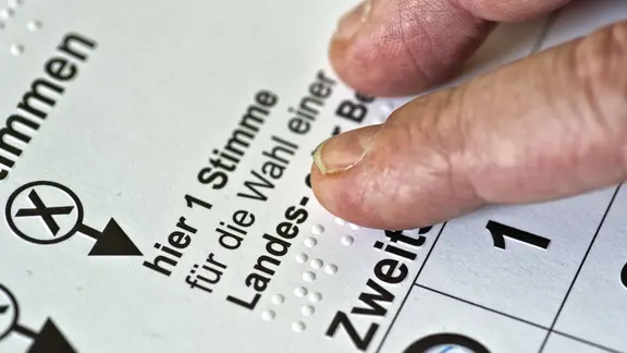 Mit einem Zeigefinger wird die Blindenschrift auf einer Wahl-Schablone gelesen.