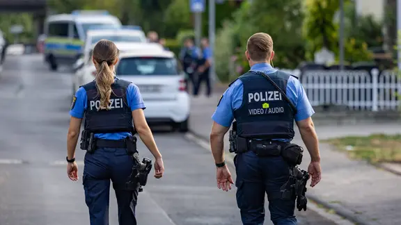 Eine Polizistin und ein Polizist laufen nebeneinander auf einer asphaltierten Straße auf weitere Kolleginnen und Kollegen zu