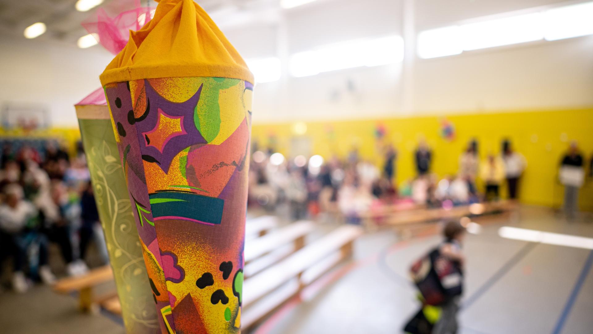 In einer Turnhalle sind Bänke, Kinder und Erwachsene im HIntergrund zu sehen. Im Vordergrund des Bildes sind zwei Schultüten abgebildet. | picture alliance/dpa | Fabian Sommer