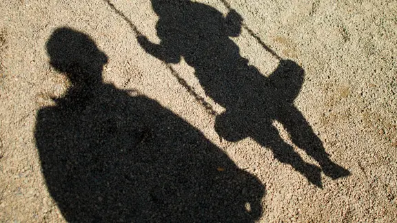 Der Schatten von einem Mann und einem schaukelnden Flüchtlingsjungen fallen auf Sand auf einem Spielplatz