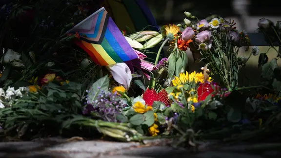 Die Sonne scheint auf Blumen und eine Progress-Pride-Fahne, die zum Gedenken an die Opfer des Anschlags auf den CSD abgelegt wurden.