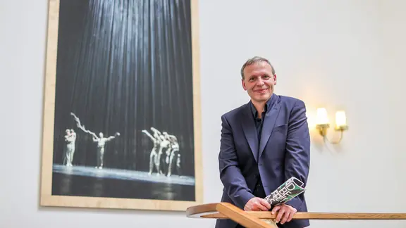 Enrico Lübbe, Intendant, steht im Schauspiel Leipzig: Ein Mann im blauen Sakko steht an einem Geländer und lächelt in die Kamera, im Hintergrund hängt ein großes Foto einer Theaterinszenierung.