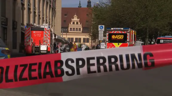 Hinter einem Polizeiabsperrband stehen mehrere Einsatzfahrzeuge. Zwischen ihnen steht eine Gruppe Menschen.