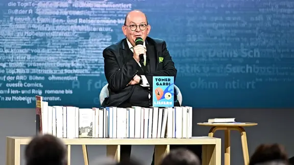 Literaturkritiker Denis Scheck bei der Frankfurter Buchmesse auf der Bühne. Vor ihm ein Tisch, auf dem eine Menge Bücher aufgereiht sind. 