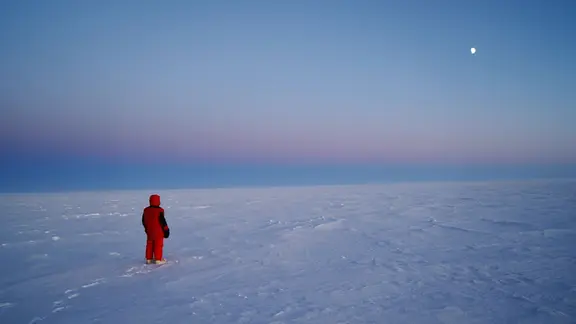 Auf einer endlosen, flachen Schneelandschaft steht eine einzelne Person in rotem Antarktis-Anzug und dicken Stiefeln, von hinten sichtbar; der Horizont teilt das Bild, darüber ein ruhiger pastellfarbener Abendhimmel mit einem kleinen, hellen Mond rechts oben. Die Szene wirkt still und weit, ohne andere Personen oder Gebäude.