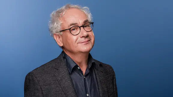 Tom Pauls, ein Mann mit Brille und grauen Haaren im Anzug, blickt verschmitzt in die Kamera.