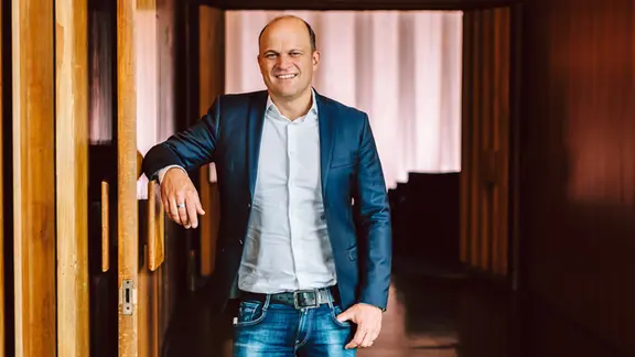 Christian Bräuer: Lächelnder Mann mit Halbglatze in blauer Jeans, hellem Hemd und dunkelblauem Jackett lehnt an einer Holztür am Eingang zu einem Kinosaal