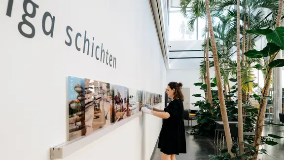 Eine Frau steht vor einer Wand, an der mehrere Fotos in einer Reihe hängen, in ihrem Rücken mehrere Pflanzen.