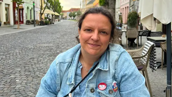 Eine Frau mit dunklen Haaren zum Zof gebunden, in hellblauer Jeansjacke sitzt an einem Cafe-Tisch mit einem roten Getränk in einer Altstadtgasse mit Pflastersteinen und lächelt in die Kamera (Stefanie Bose in Trenčín)