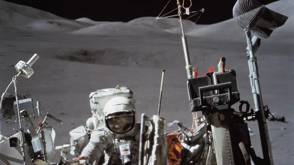 Astronaut Harrison Schmitt auf dem Mond. Vor ihm steht der Mondrover, hinter ihm ist eine karge Gegend mit Mondstaub zu sehen.