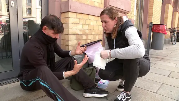 Eine junge Frau hockt bei einem jungen Mann, der in einem Fußgängerbereich auf dem Boden sitzt und ihr beide Hände entgegenhält. In ihren Händen hält sie ein großes Wundpflaster.