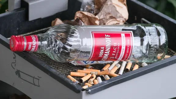 Im Aschenbecher eines Mülleimers liegen mehrere Zigarettenkippen, eine leere Wodka-Flasche und eine zusammengeknüllte Bäckertüte