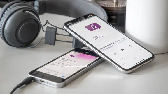 Zwei Mobiltelefone mit einem MDR KLASSIK-Podcast. Dahinter Kopfhörer, eine Kaffeetasse und eine French Press als Kaffeekanne.