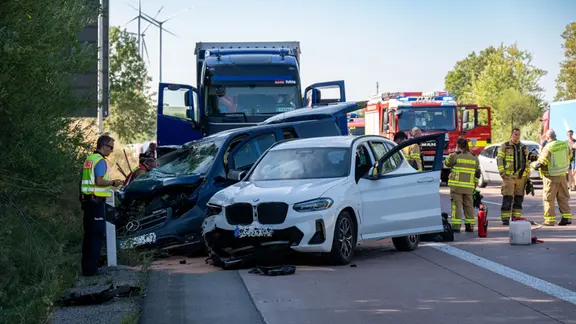 Vor einem LKW auf der Autobahn stehen zwei kollidierte Fahrzeuge schräg zur Fahrtrichtung. Daneben stehen Einsatzkräfte der Feuerwehr. 