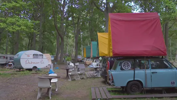 Auf einem Campingplatz stehen Trabanten mit Zelten auf dem Dach und ein Dübener Ei.
