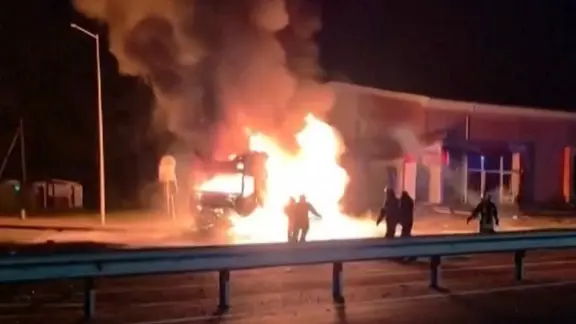 Ein brennendes Fahrzeug vor einem Gebäude in der Nacht. Davor fünf Menschen.