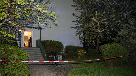 Weißrote Bänder sperren den Eingang zu einem Eingang in einem Plattenbau ab. Im Eingang steht ein unformierter Polizist. 