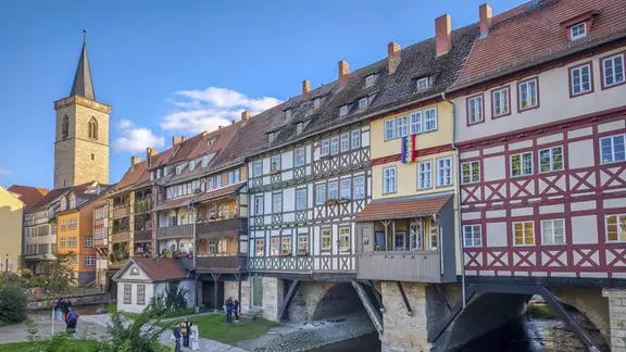 Mittelalterliche Nordseite der gotischen Krämerbrücke mit geschlossener Brückenbebauung mit Fachwerkhäusern und darunter das Krämerbrückengewölbe über dem Fluss Gera, ganz links sieht man den steinernen Kirchenturm in der Altstadt von Erfurt