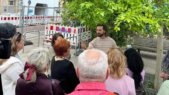 Im Freien vor einem Baum und einem Haus, vor dem ein Baustellenzaun steht, steht eine Gruppe Menschen einem anderen erwachsenen Menschen zugewandt