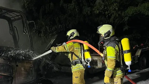 Zwei Feuerwehrleute sprühen Löschwasser auf einen ausgebrannten Mini-Bagger.