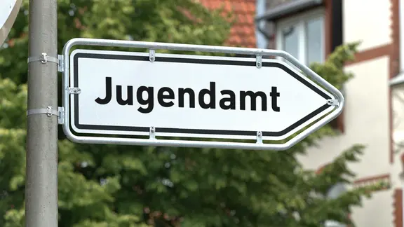 Ein Schild auf dem "Jugendamt" steht, dahinter ein Haus und ein Baum