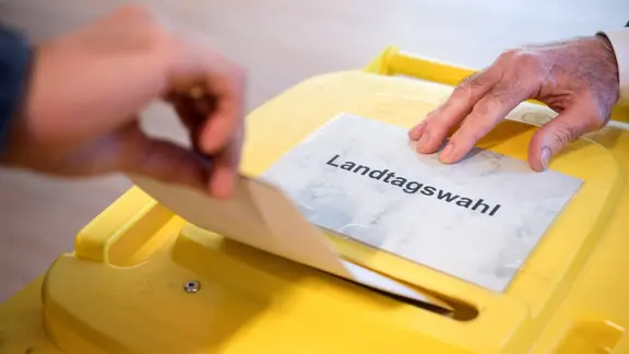 Eine gelbe Wahlurne, auf der ein Zettel mit dem Wort Landtags-Wahl liegt. 