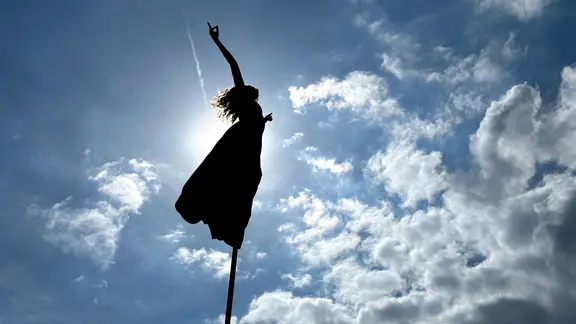 Szene aus der Open-Air-Performance "RoZéO"  der Kompanie Gratte Ciel: Eine Artistin schwebt auf einer Stange hoch in der Luft. Das Bild ist von unten aufgenommen, so das es aussihet, als würde sie in die Wolken fliegen