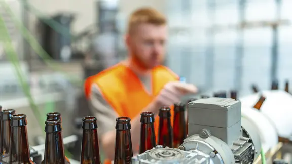 Braunglasflaschen, die sich auf einem Förderband bewegen, mit einem Fabrikarbeiter in einer orangefarbenen Weste, der den automatisierten Abfüllprozess in einer Bierproduktionsanlage überwacht.